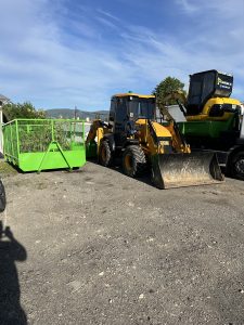 Traktorbager JCB 3CX Compact a bager CAT zaparkované vedľa zeleného kontajnera. Stroje spoločnosti po-bagrujem.sk pripravené na výkopové práce, zemné úpravy a prepravu stavebného materiálu. Ideálne na realizáciu výkopových prác a zemných prác na rôznych typoch stavieb a projektov.